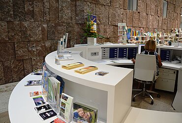 Auslage von Flyern am Informationszentrum. Im Hintergrund der Buchladen mit niedrigen weißen Regalen und vielen Büchern